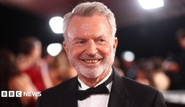 Graham Norton has white hair, a moustache and beard. He is wearing a dark-colour T-shirt and sits behind a microphone with a green muffler.