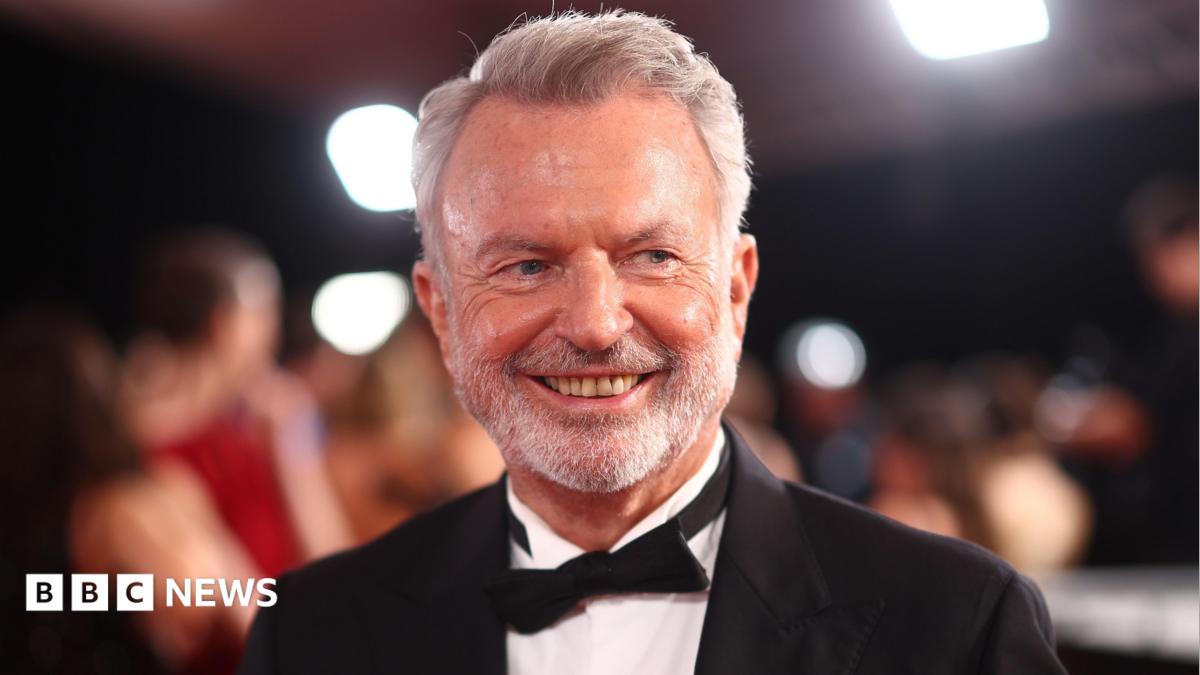 Graham Norton has white hair, a moustache and beard. He is wearing a dark-colour T-shirt and sits behind a microphone with a green muffler.