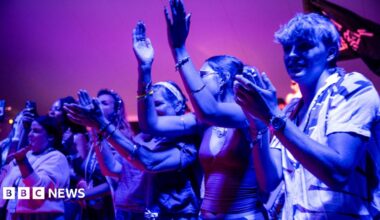 A group of several young people, illuminated by blueish light, applaud as they watch a live performance at WOMAD 2026