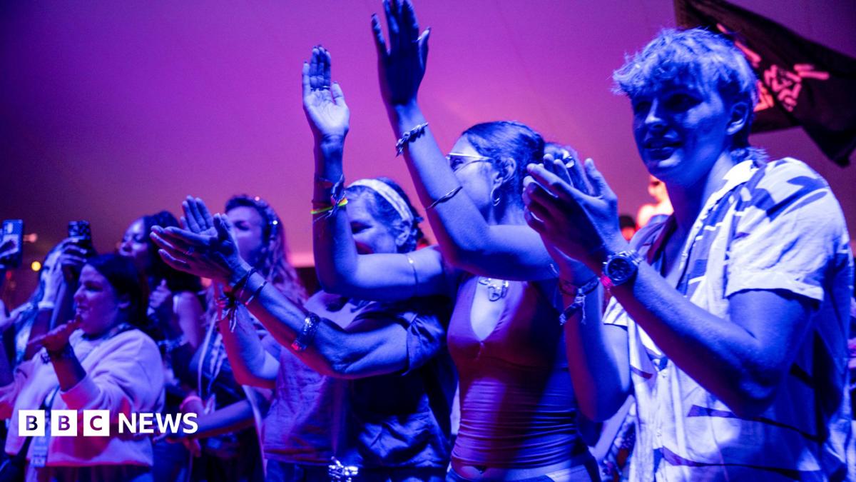 A group of several young people, illuminated by blueish light, applaud as they watch a live performance at WOMAD 2026