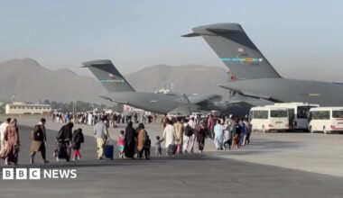 People carrying luggage walk across an airport tarmac towards large US military transport aircraft during an evacuation