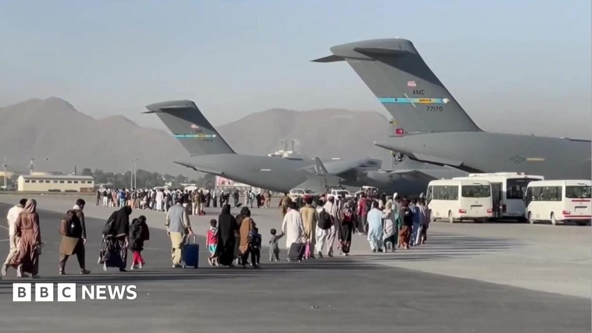 People carrying luggage walk across an airport tarmac towards large US military transport aircraft during an evacuation