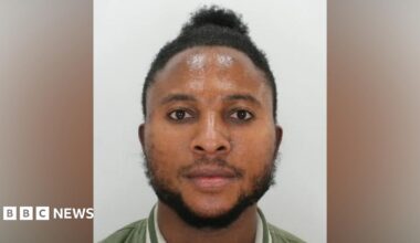 A police custody image of a man. He has short black hair, a black beard and is wearing a green jacket with white stripes around the collar.