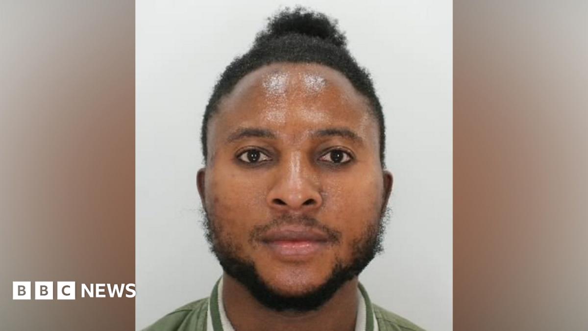 A police custody image of a man. He has short black hair, a black beard and is wearing a green jacket with white stripes around the collar.