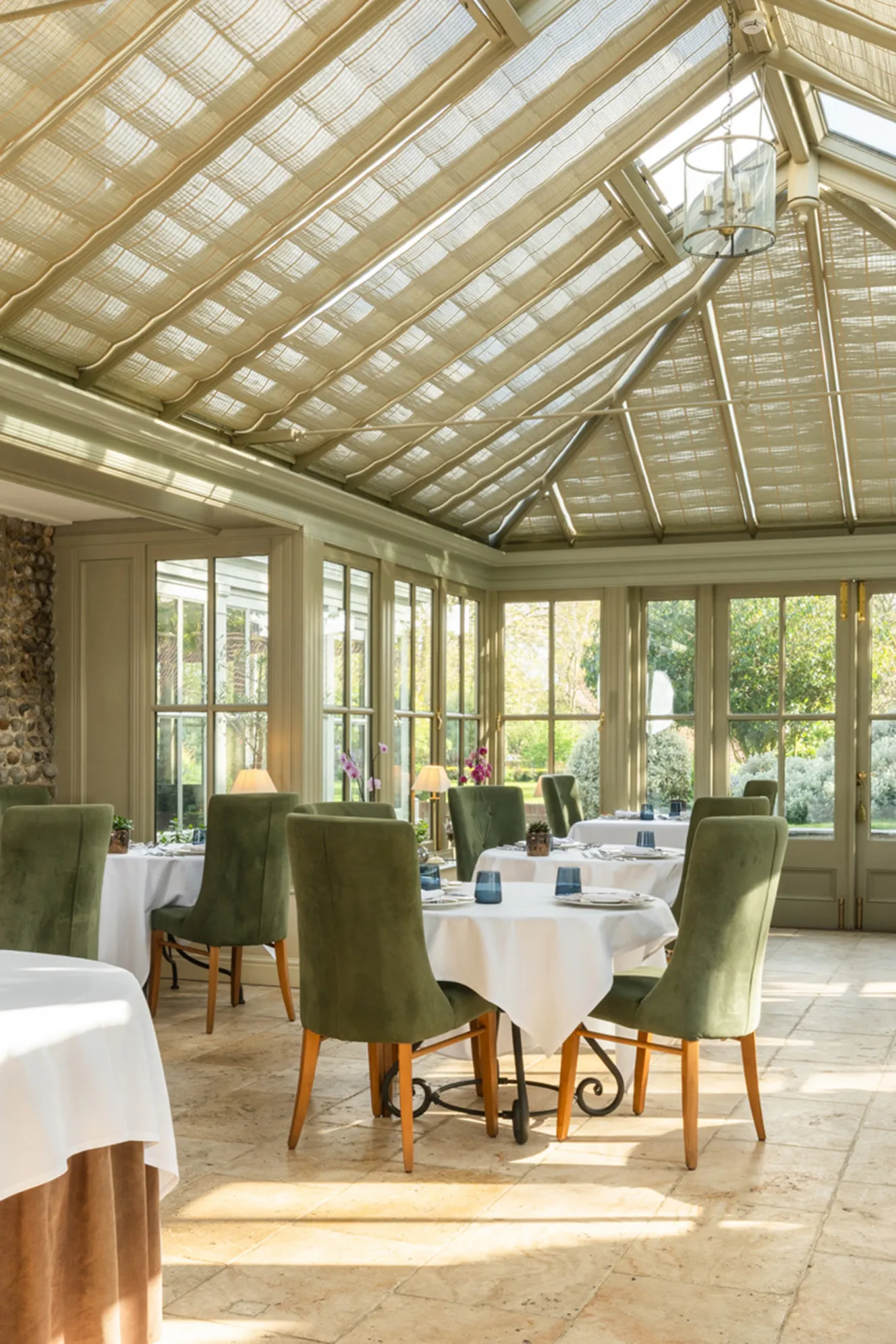 The sunlit dining room at Morston Hall, featuring tables set with white tablecloths and dark green chairs, under a glass ceiling with blinds.