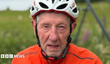 Eddie looks directly at the camera with a determined look. He's wearing a white cycling helmet and orange top.