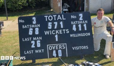 A man in cricket whites knelt next to a cricket scoreboard.