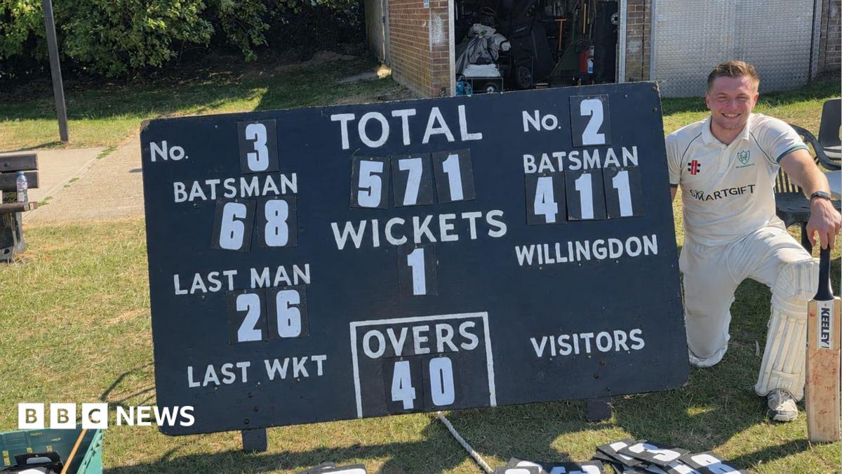 A man in cricket whites knelt next to a cricket scoreboard.