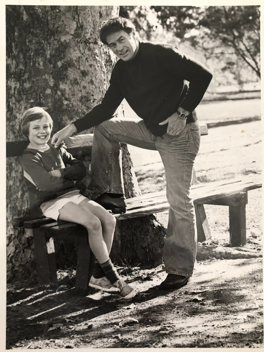 A black and white photo of a young boy sitting on a park bench and a man standing beside him with one foot on the seat.