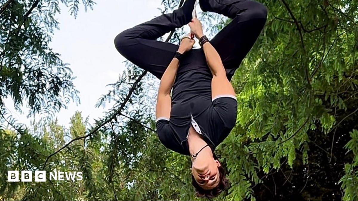 A man hangs upside from a rope with his legs apart at the knees. He's looking down. Behind him are trees and a blue sky.