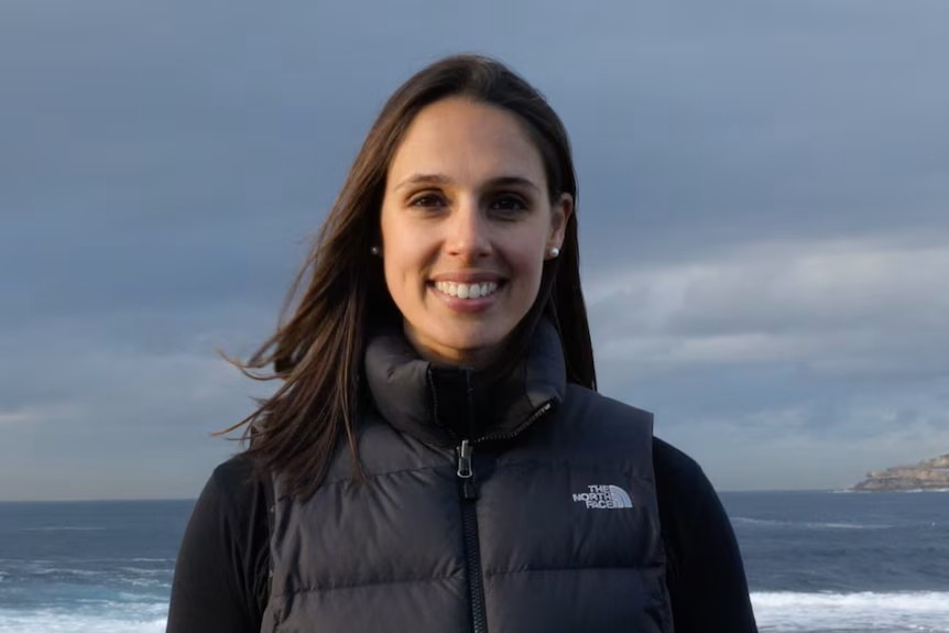 A brunette woman standing in front of the ocean waring a black puffer jacket.