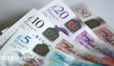 Four banknotes are in a pile on a table. Each banknote is partially obscured. There is a £50 note in the background which has a £20 on top of it. A £10 is on top of the £20, and a £5 is on top of the £10.