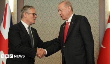 Sir Keir and Erdogan, both wearing dark suits, shake hands. Behind them are the Union Jack and Turkish national flag.