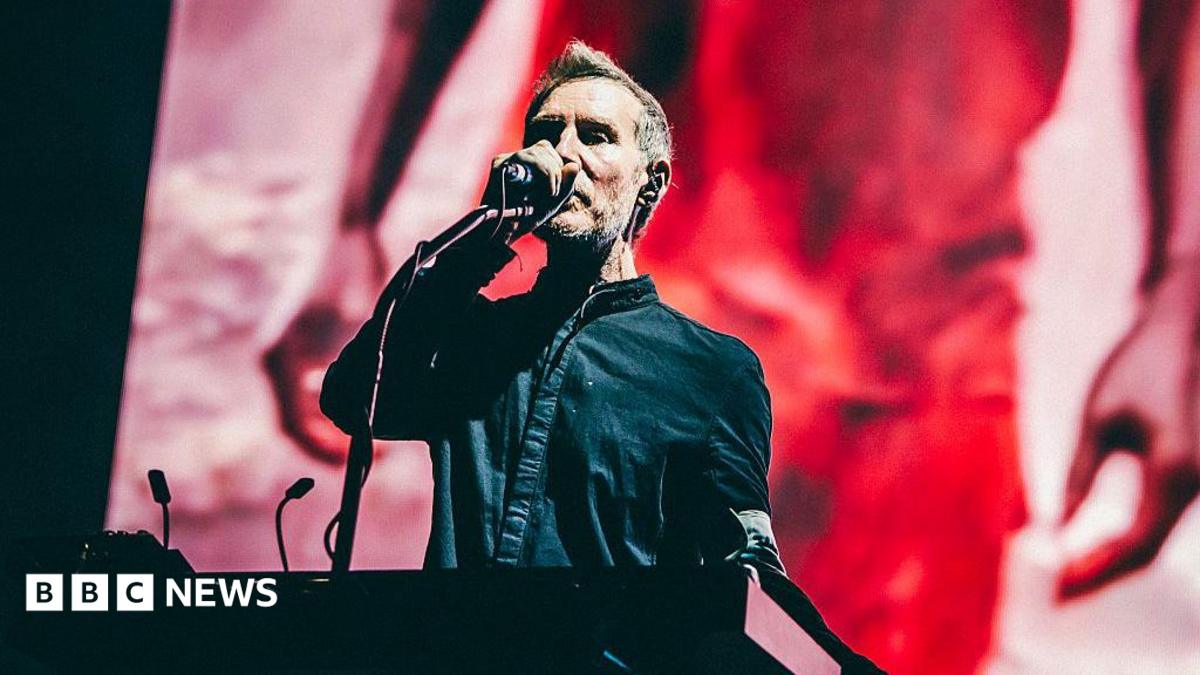 Robert Del Naja, one of the band members of Massive Attack, sings into a microphone while standing on stage at a keyboard