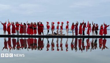 A long line of women wearing red dresses photographed standing next to a lake, with their reflection immediately below in the water.