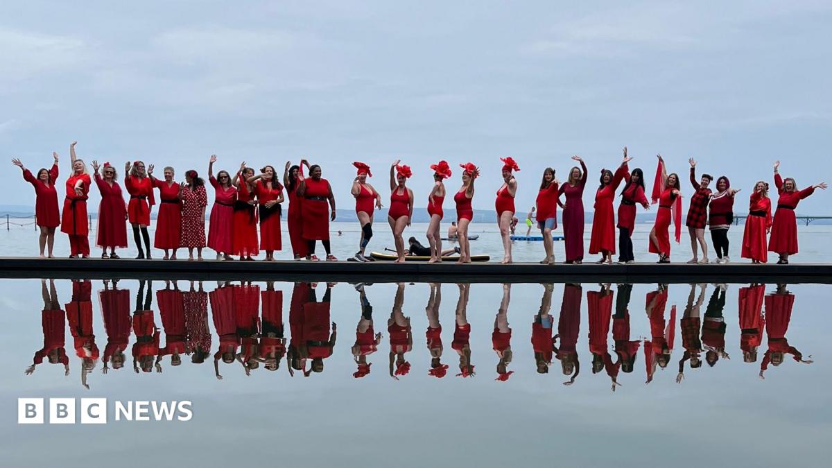 A long line of women wearing red dresses photographed standing next to a lake, with their reflection immediately below in the water.