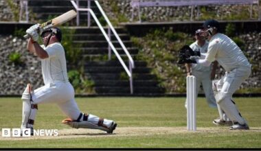 A batsman stretches out of his crease to hit a scoring shot. A wicket keeper stands behind the stumps. Both men are in full whites cricket gear. Another fielder is seen in the background. The pitch they are playing on is well maintained.