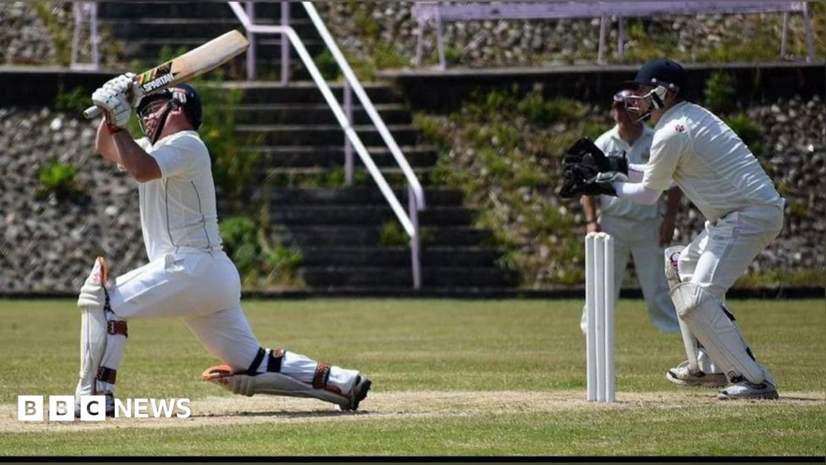 A batsman stretches out of his crease to hit a scoring shot. A wicket keeper stands behind the stumps. Both men are in full whites cricket gear. Another fielder is seen in the background. The pitch they are playing on is well maintained.