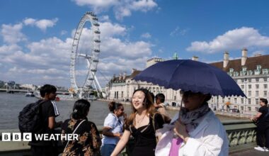 People walk on a bridge over the Thames in London using an umbrella for shade with strong sunshine overhead.