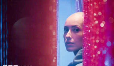 Emily Glaze is turning to look at the camera side-on from between pink glittery curtains. She has a bald head and is wearing a light blue hoodie.
