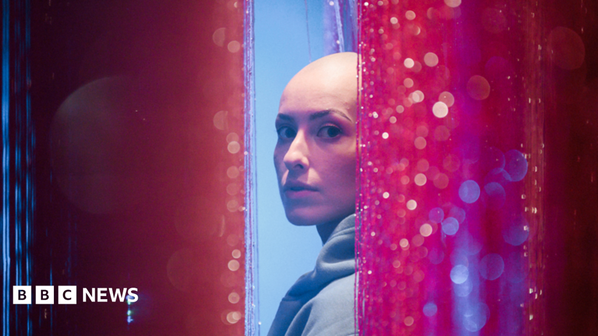 Emily Glaze is turning to look at the camera side-on from between pink glittery curtains. She has a bald head and is wearing a light blue hoodie.