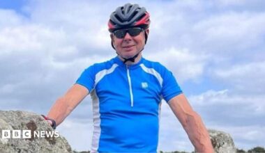 John Lyttle wearing a blue and white short-sleeved athletic top, sunglasses and a grey bicycle helmet. He is standing outdoors on a sunny day, with lots of fluffy clouds behind him, leaning against a rock covered in lichen and smiling at the camera.