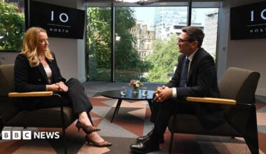 Andy Burnham, wearing a dark suit, gestures with his hands while speaking during an interview. Behind him is a TV with the logo for No 10 North.