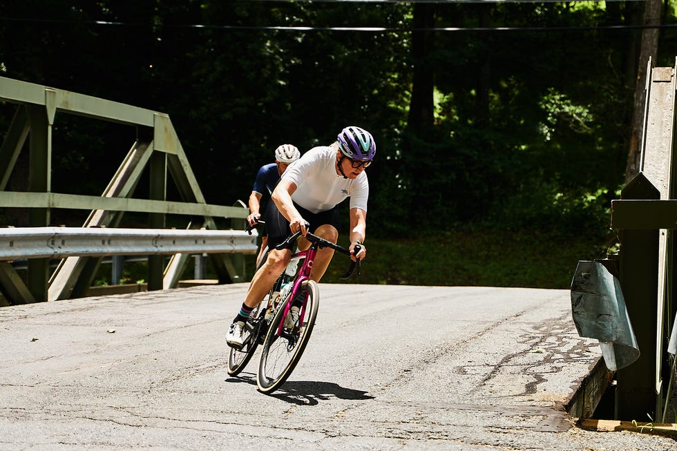 Two cyclists riding over a bridge on a sunny day, one leading with a focused expression.