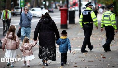 A family including two young girls in white dresses and pink coats along with a little boy in a blue coat. They walk down the street with their backs to the camera and in the background two Metropolitan Police officers in uniform can be seen walking in the same direction.