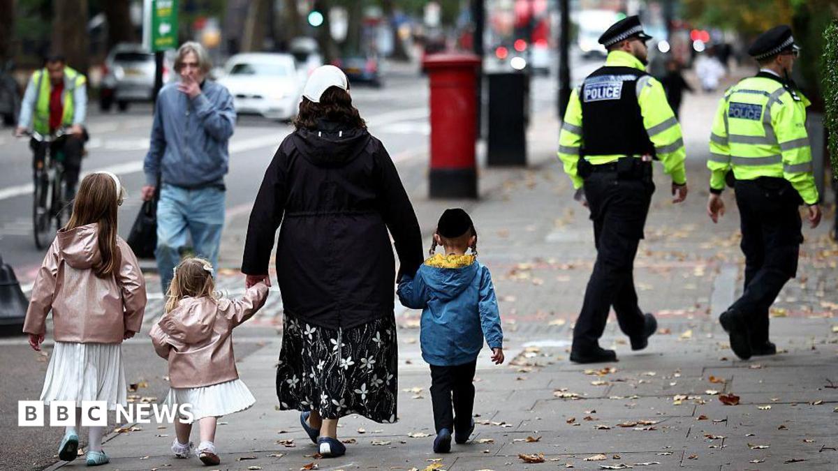 A family including two young girls in white dresses and pink coats along with a little boy in a blue coat. They walk down the street with their backs to the camera and in the background two Metropolitan Police officers in uniform can be seen walking in the same direction.