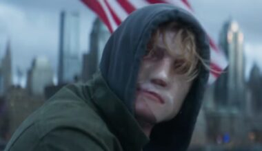 closeup of man in a dark hoody sitting outside, his disfigured face covered by a mask