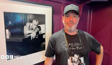 Steve Read stands beside a framed black-and-white photograph. Read is wearing a dark baseball cap and a black T-shirt featuring artwork from the exhibition. The photograph beside him shows a blond-haired musician leaning on a counter, while burgundy-coloured wall panels form the background.