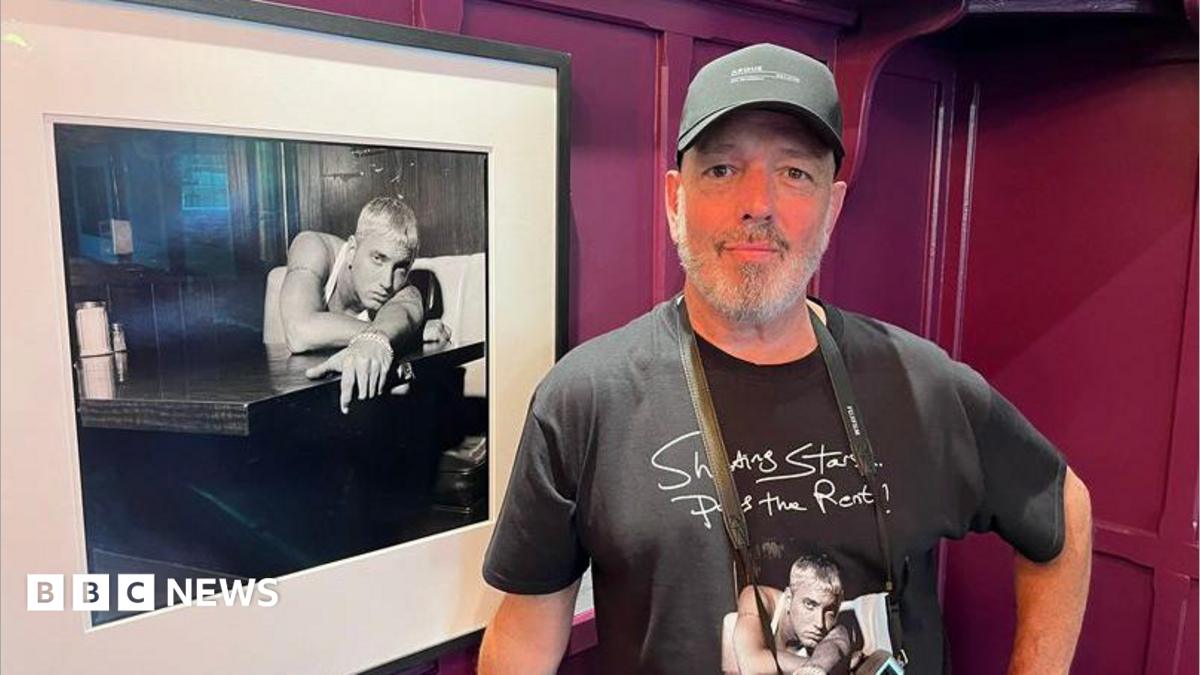 Steve Read stands beside a framed black-and-white photograph. Read is wearing a dark baseball cap and a black T-shirt featuring artwork from the exhibition. The photograph beside him shows a blond-haired musician leaning on a counter, while burgundy-coloured wall panels form the background.