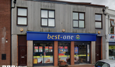 A shop front. The shop is the ground floor of a brick building with wood over the bricks. It has blue hoarding above with best written in yellow and one written in while.