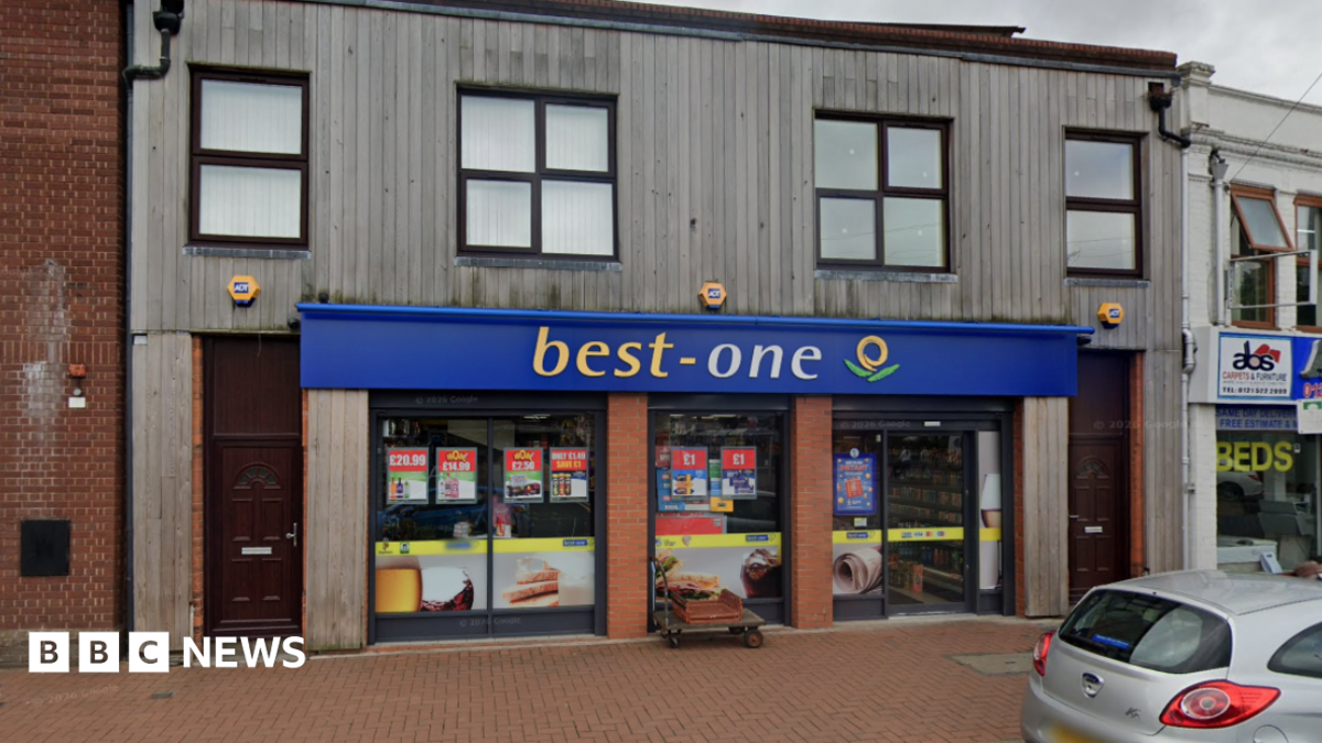 A shop front. The shop is the ground floor of a brick building with wood over the bricks. It has blue hoarding above with best written in yellow and one written in while.