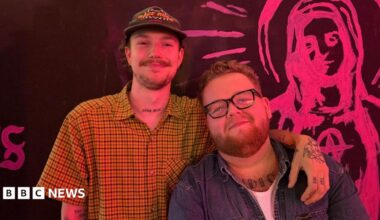 Two men pose together in front of a black wall featuring pink artwork. On the left, a man wears a yellow checked shirt and cap; on the right, a man wears glasses and a denim shirt.