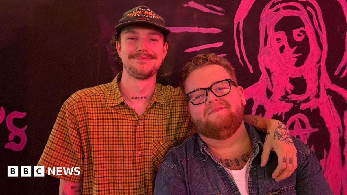 Two men pose together in front of a black wall featuring pink artwork. On the left, a man wears a yellow checked shirt and cap; on the right, a man wears glasses and a denim shirt.