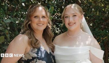 Hannah and her mother at Hannah's wedding. Her mother is wearing a blue dress with white flowers. Hannah on the right is dressed in a white wedding dress holding a bouquet