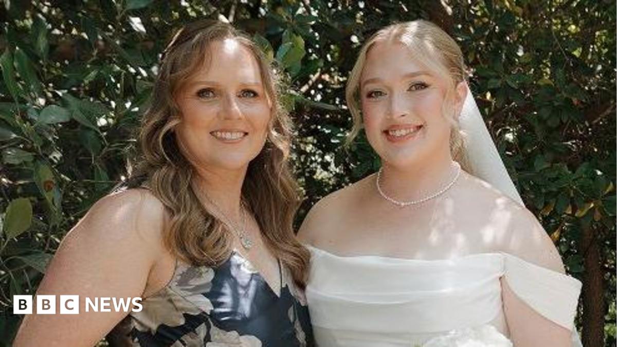 Hannah and her mother at Hannah's wedding. Her mother is wearing a blue dress with white flowers. Hannah on the right is dressed in a white wedding dress holding a bouquet