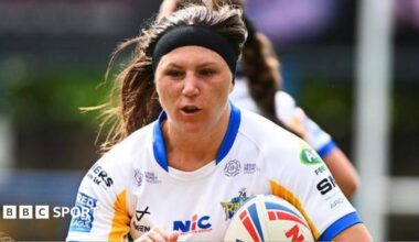 Leeds Rhinos player Connie Boyd, in white, blue and gold and wearing a hairband, carries the ball.