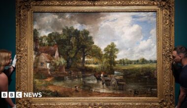A large landscape oil painting in an ornate gilded frame is held in position on a wall by a woman to the left and a man to the right. The painting is The Hay Wain by John Constable, from 1821, and depicts a horse and cart in a shallow river, with a white house to the left. The scene is surrounded by trees and fields.