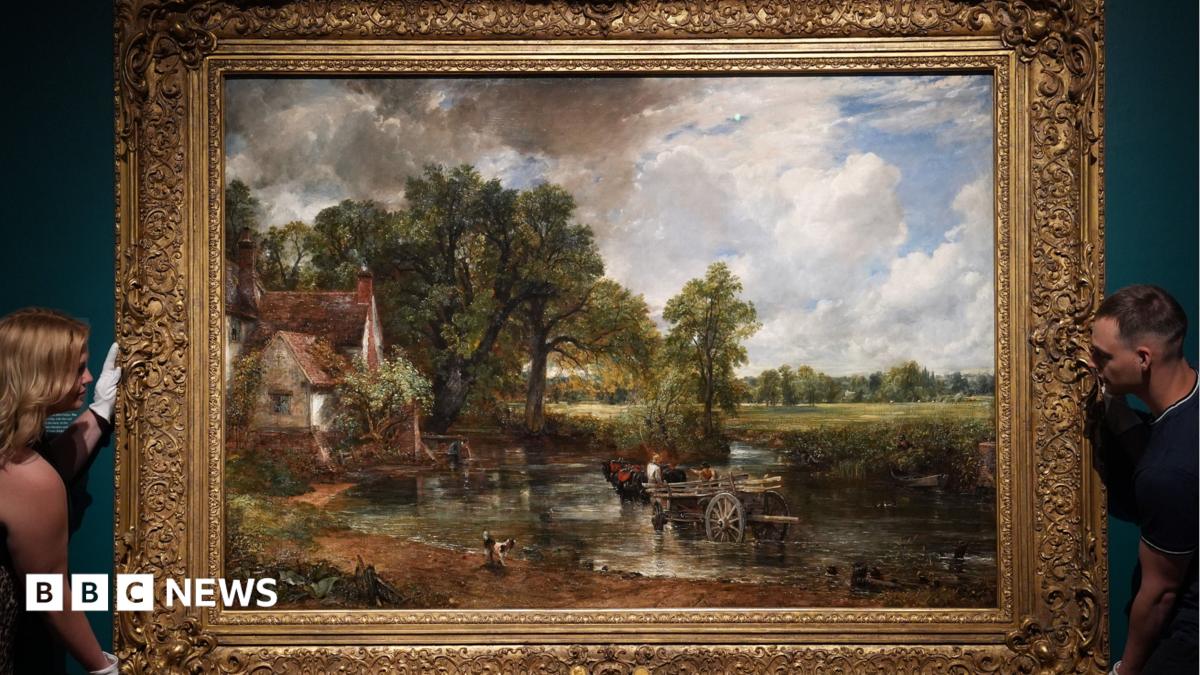 A large landscape oil painting in an ornate gilded frame is held in position on a wall by a woman to the left and a man to the right. The painting is The Hay Wain by John Constable, from 1821, and depicts a horse and cart in a shallow river, with a white house to the left. The scene is surrounded by trees and fields.