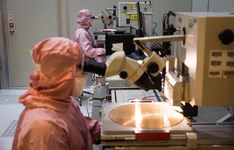 Two scientists in full light pink cleanroom suits operating high-powered microscopes to inspect large circular silicon wafers used in semiconductor microchip fabrication.