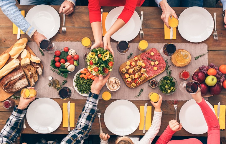 A top-down view of a festive dining table set for six people with empty white plates, yellow napkins, and various dishes including salad, bread, charcuterie, and fruit. Hands of six individuals reach in to serve or hold glasses of orange juice and red wine.
