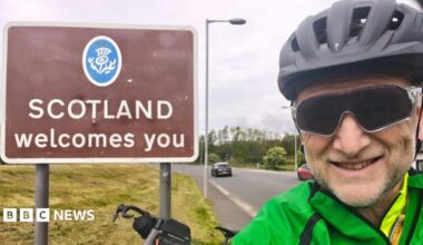 A picture of Graham Beck wearing a green jacket. He is standing in front of a brown sign that says: Scotland welcomes you. He is wearing black glasses and a helmet.