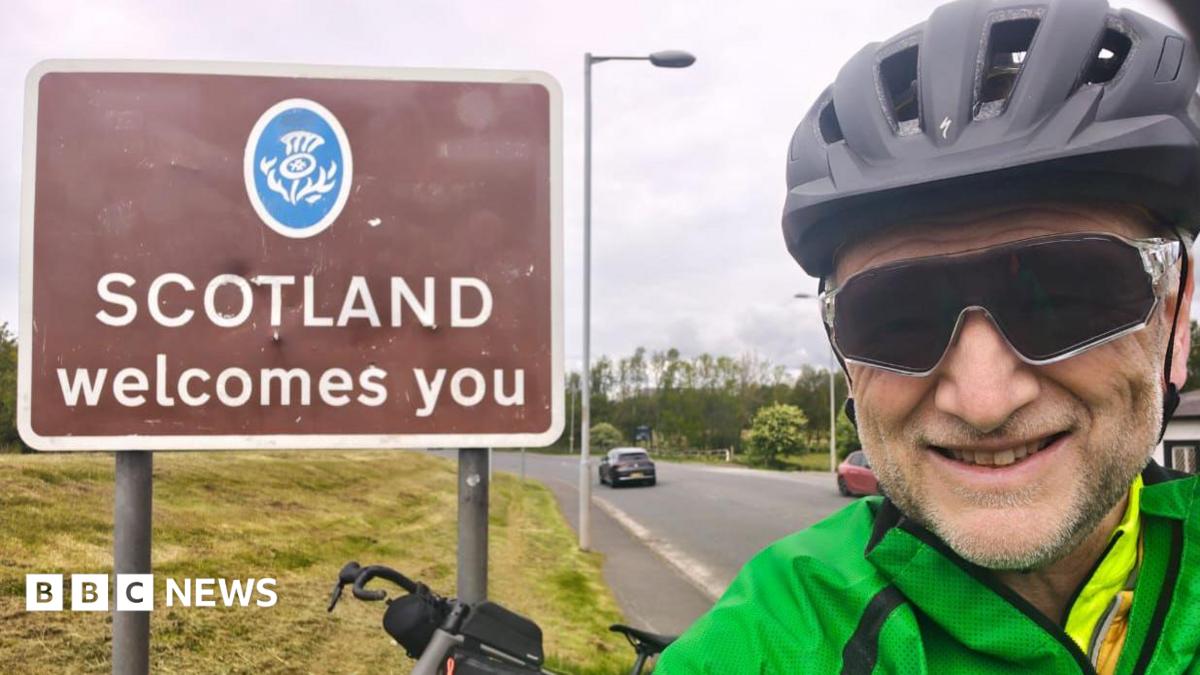 A picture of Graham Beck wearing a green jacket. He is standing in front of a brown sign that says: Scotland welcomes you. He is wearing black glasses and a helmet.