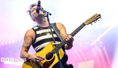 Scottish singer-songwriter Callum Beattie is performing on a stage. He is wearing a black and white stripy vest and is playing a yellow guitar.
