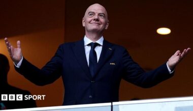 Gianni Infantino holds his arms out at each side with open palms, as he smiles while wearing a buttoned-up, dark smart jacket with FIFA written on its breast pocket