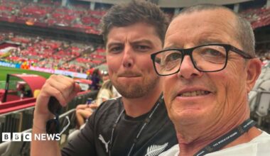 Two football fans in a stadium. The one nearest the camera has dark/grey receding short hair and glasses. The one behind his is younger nad has dark spiky hair and it in a black New Zealand top and is holding sunglasses in his right hand.