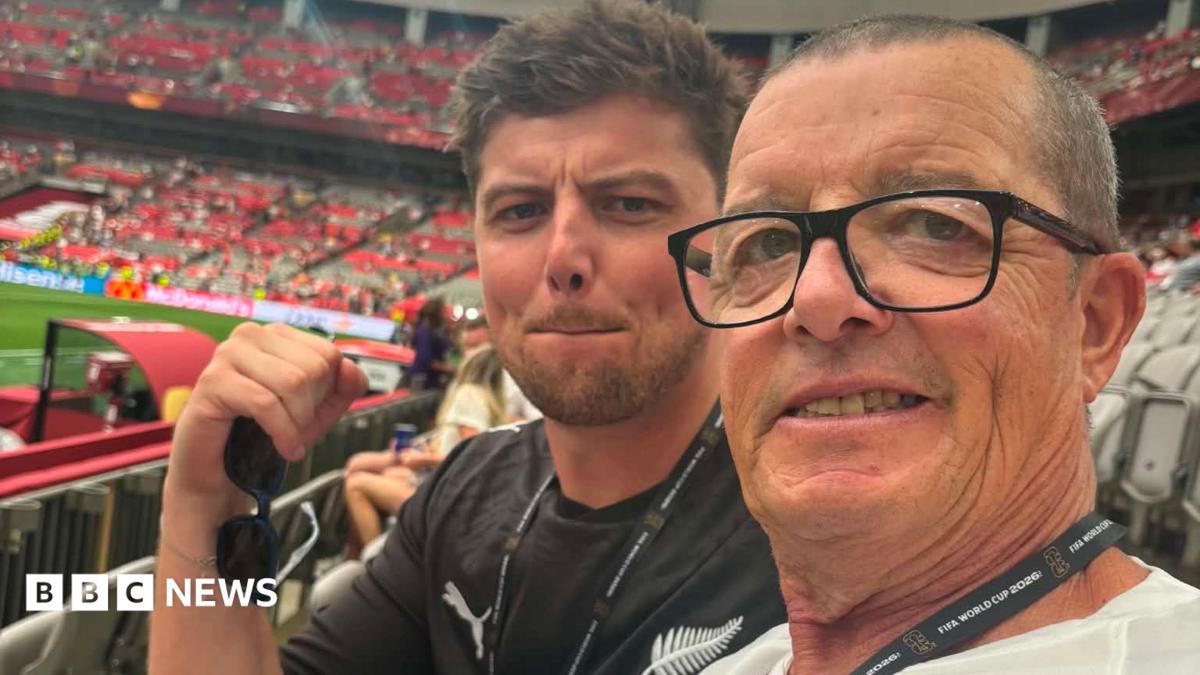 Two football fans in a stadium. The one nearest the camera has dark/grey receding short hair and glasses. The one behind his is younger nad has dark spiky hair and it in a black New Zealand top and is holding sunglasses in his right hand.